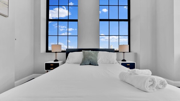 A large bed with white linens, two bedside tables with lamps, a gray pillow, and a view through a large window.
