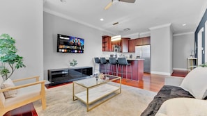 A modern living room with a flat-screen TV, a sofa, a coffee table, and a kitchen area with a refrigerator and bar stools.