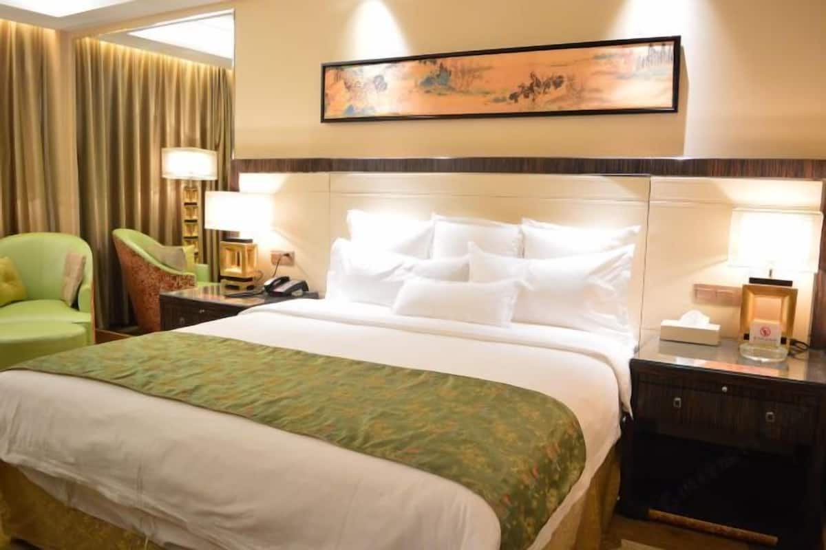 Une chambre d’hôtel avec un grand lit, un fauteuil vert, un bureau avec une lampe et un tableau au mur.