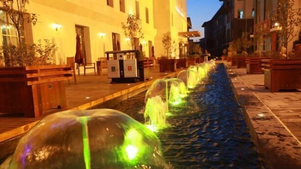 Un espace extérieur aménagé pour la nuit, avec des fontaines illuminées, des bancs et des plantes en pot, le tout disposé le long d’un chemin pavé.