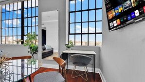 A modern living room with large windows, a grey sofa, a small table, and a TV displaying streaming services.