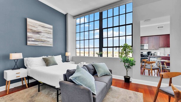 A modern bedroom with a large bed, bedside tables, a sofa, a dining area, and a view of the city through a large window.