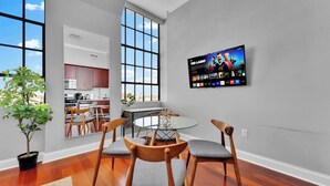 A modern kitchen with wooden cabinets, a dining area with a glass table and chairs, and a TV displaying a sports channel.