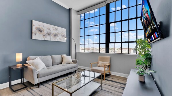 A modern living room with a grey sofa, a coffee table, a side table with a lamp, a wall-mounted TV, and a large window with a city view.