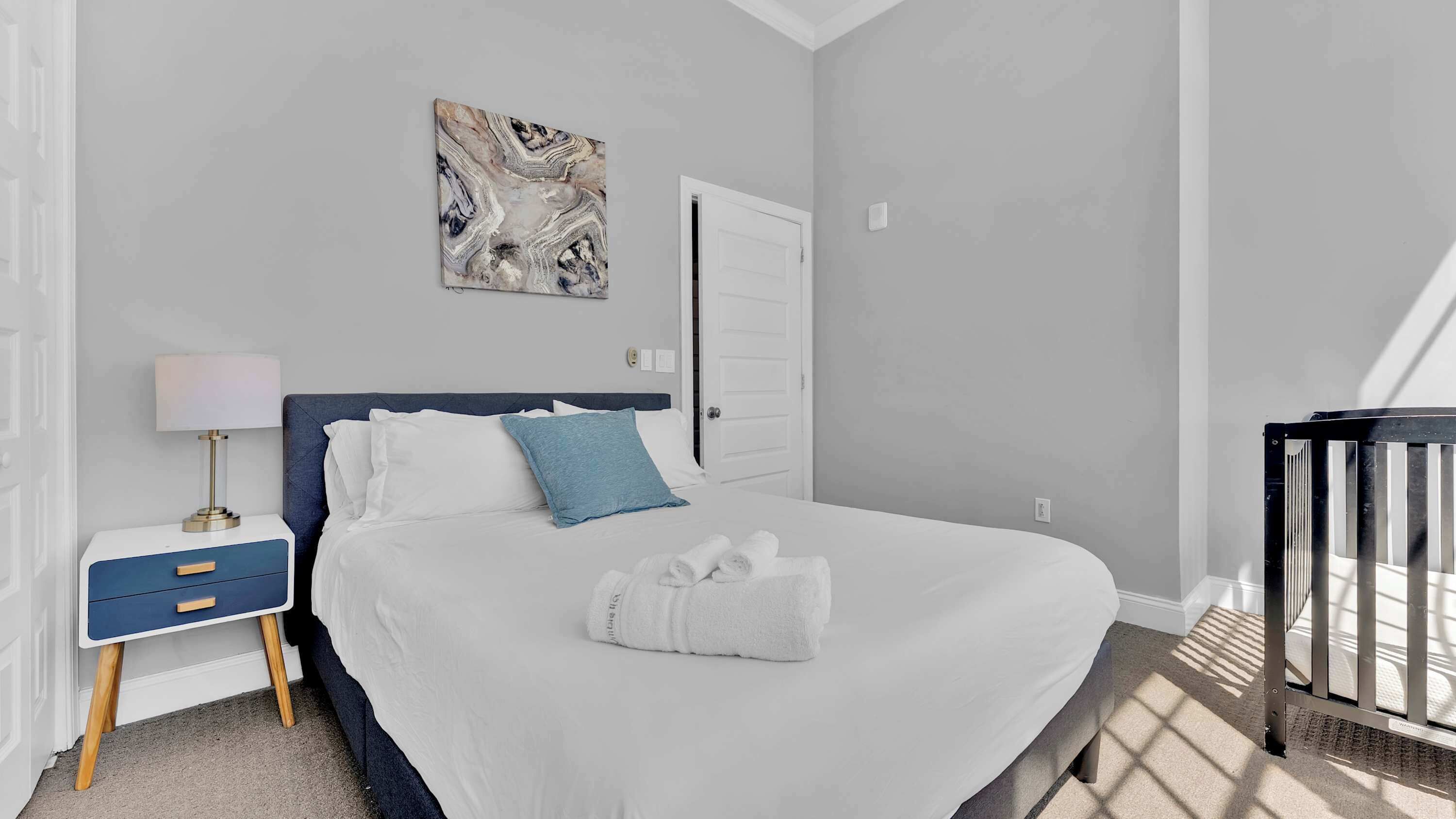 A bedroom with a bed, a bedside table with a lamp, a door, and a wall-mounted artwork.