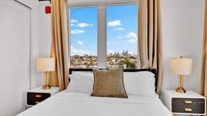 Quarto de hotel com cama, duas mesas de cabeceira com luminárias, janela com cortinas e vista para a cidade.