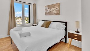 A hotel room with a large bed, a nightstand, a lamp, a painting, and a window with curtains.