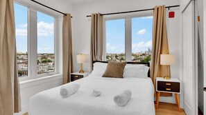 A hotel room with a large bed, two bedside tables, a view of the city, and curtains.