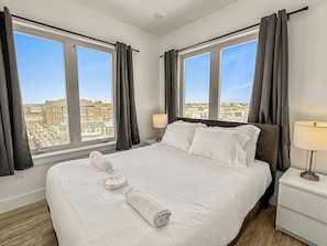 A hotel room with a large bed, two bedside tables, and a view of the cityscape through large windows. - Comfy 2BR for 4 | Philly Getaway Near Philly Spots (Philadelphia)