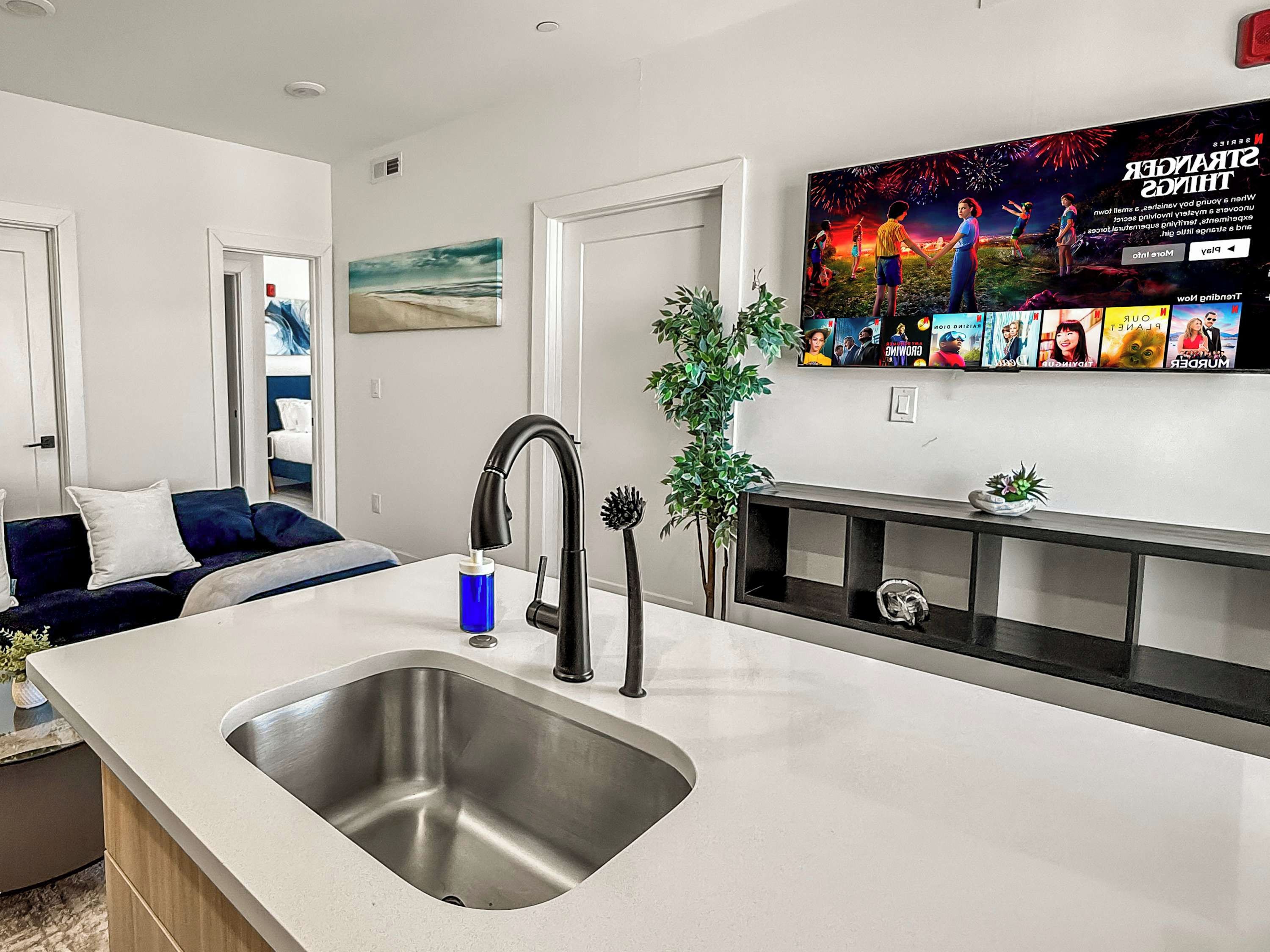 A modern kitchen with a stainless steel sink, a black faucet, and a flat-screen TV mounted above the counter displaying a movie.