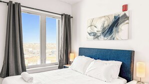 A hotel room with a large bed, two bedside tables with lamps, a view of the city through the window, and a piece of abstract art on the wall. - Cozy 2BD Apt | Fishtown | Gym (Philadelphia)