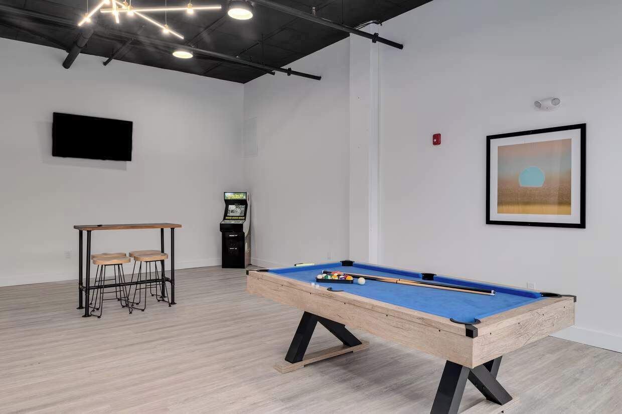 Une table de billard dans une pièce au plancher en bois, une petite table avec des tabourets, un téléviseur fixé au mur et un tableau encadré accroché au mur.