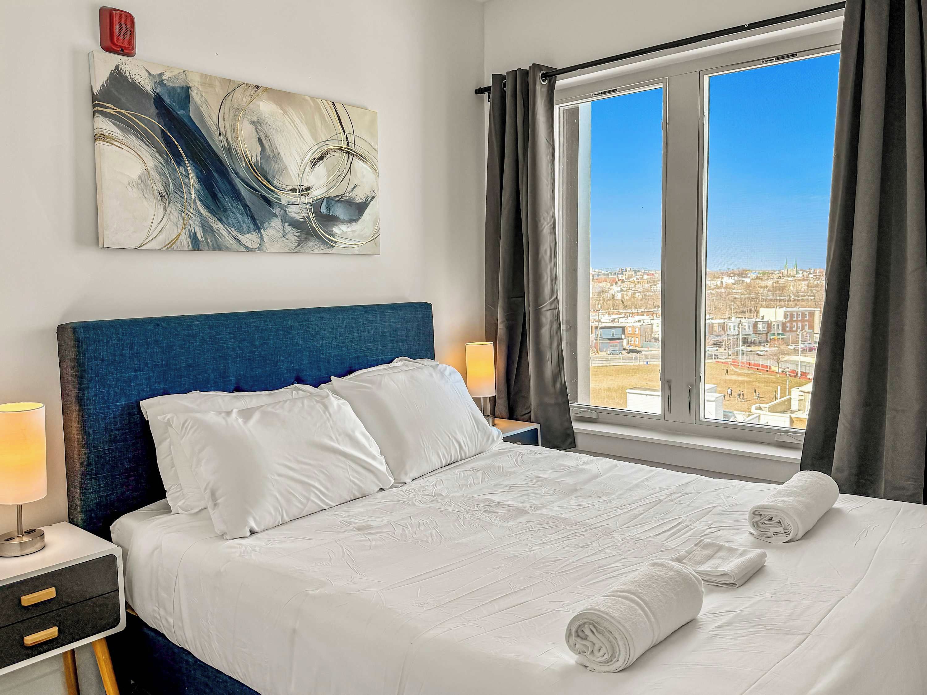 Een hotelkamer met een groot bed, een hoofdbord, een raam met gordijnen, een bedlampje en een schilderij aan de muur.