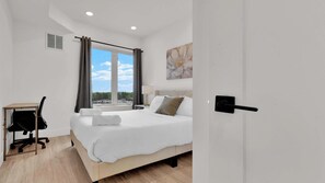 A bedroom with a bed, a desk, a chair, a window with curtains, and a door.
