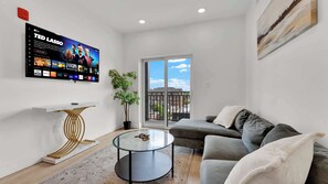 A modern living room with a flat-screen TV, a round glass coffee table, a grey sofa, and a potted plant.