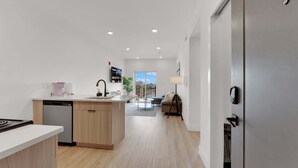 A modern kitchen with a dishwasher, a sink, and a coffee machine. There is a dining area with a table and chairs, and a view of the city through a large window. - Cozy 1BD Apt | Fishtown | Gym (Philadelphia)