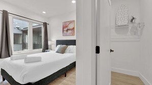 Ein Hotelzimmer mit einem Bett, einem Nachttisch, einem Fenster mit Vorhängen, einem Gemälde und einer weißen Tür.