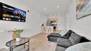 A modern living room with a flat-screen TV, a glass coffee table, a sofa, and a kitchen in the background. - Charming 1BR Home | Fishtown | Steps to Cafe & Gym (Philadelphia)