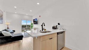 A modern kitchen with a white countertop, a sink, and a dishwasher. There is a dining area with a black sofa and a small table. A TV is mounted on the wall, and a balcony with a view of a parking lot is visible. - Charming 1BR Home | Fishtown | Steps to Cafe & Gym (Philadelphia)