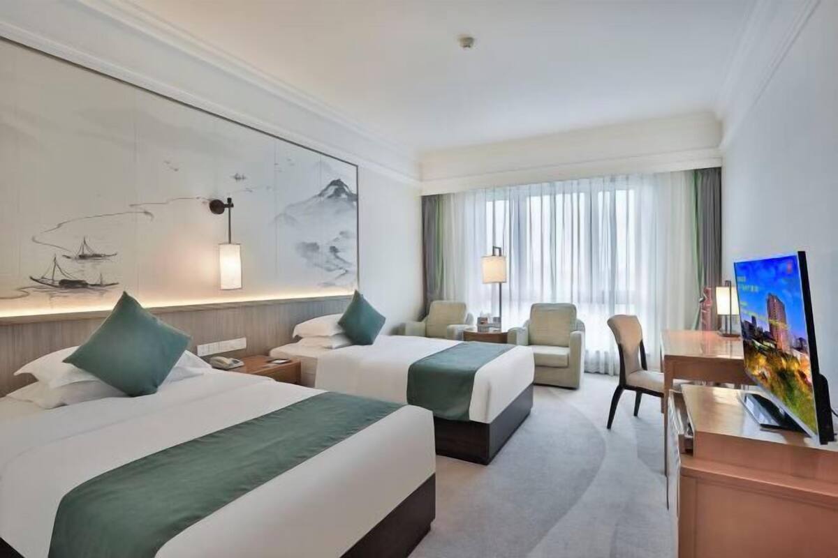 A hotel room with two beds, a desk, a chair, a TV, and a large window with curtains.