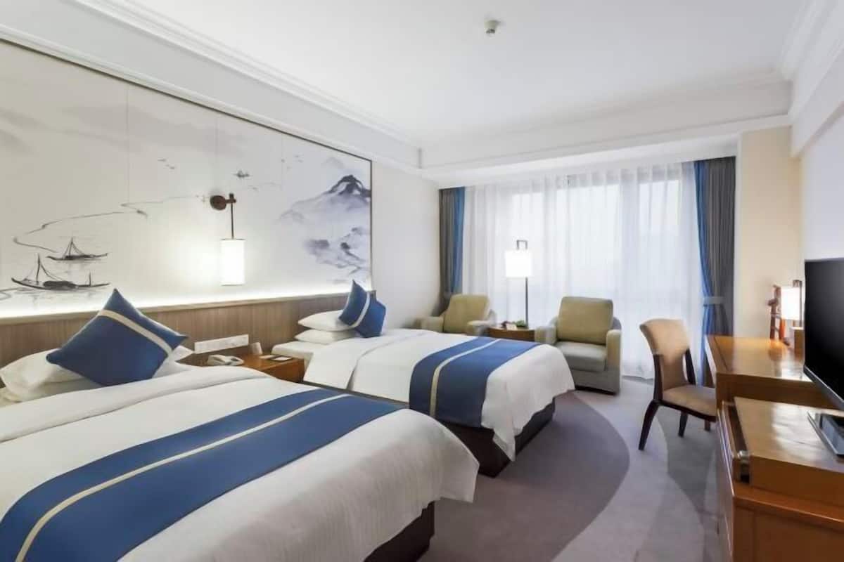A hotel room with two beds, a desk, a chair, a TV, and a large window with curtains.