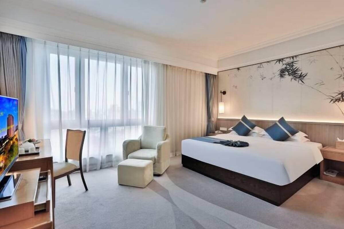 A hotel room with a large bed, a desk with a TV, a chair, and a large window with curtains.