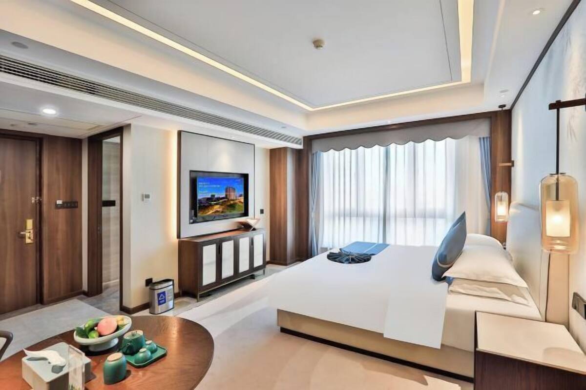 A modern hotel room with a large bed, a flat-screen TV mounted on the wall, a bedside table, and a small seating area with a table and chairs.