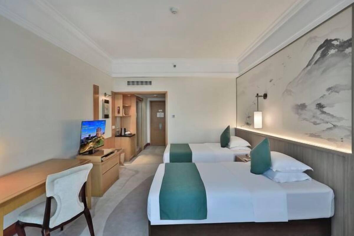 A hotel room with a desk, two beds, a TV, and a large painting on the wall.