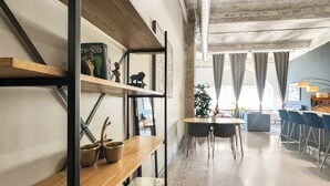 A modern interior with exposed concrete ceilings, wooden shelves, and a dining area with blue chairs. - Elegant 2BD | DT Dallas | Sleeps 10 | ValetParking (Dallas)
