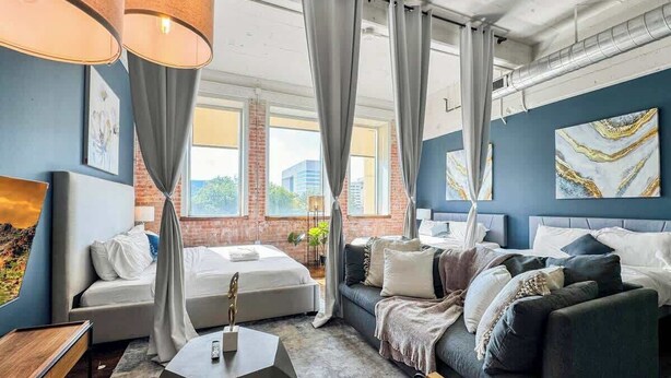 Une chambre d’hôtel moderne avec un grand lit, un canapé et une table basse. De grandes fenêtres sont agrémentées de rideaux, un tableau orne le mur et la chambre offre une vue sur la ville.