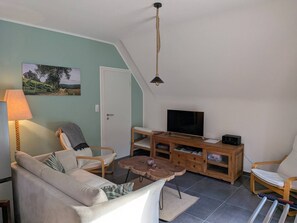 Ein Wohnzimmer mit einer Couch, Sesseln, einem Fernseher, einer Holzkommode und einer Lampe.
