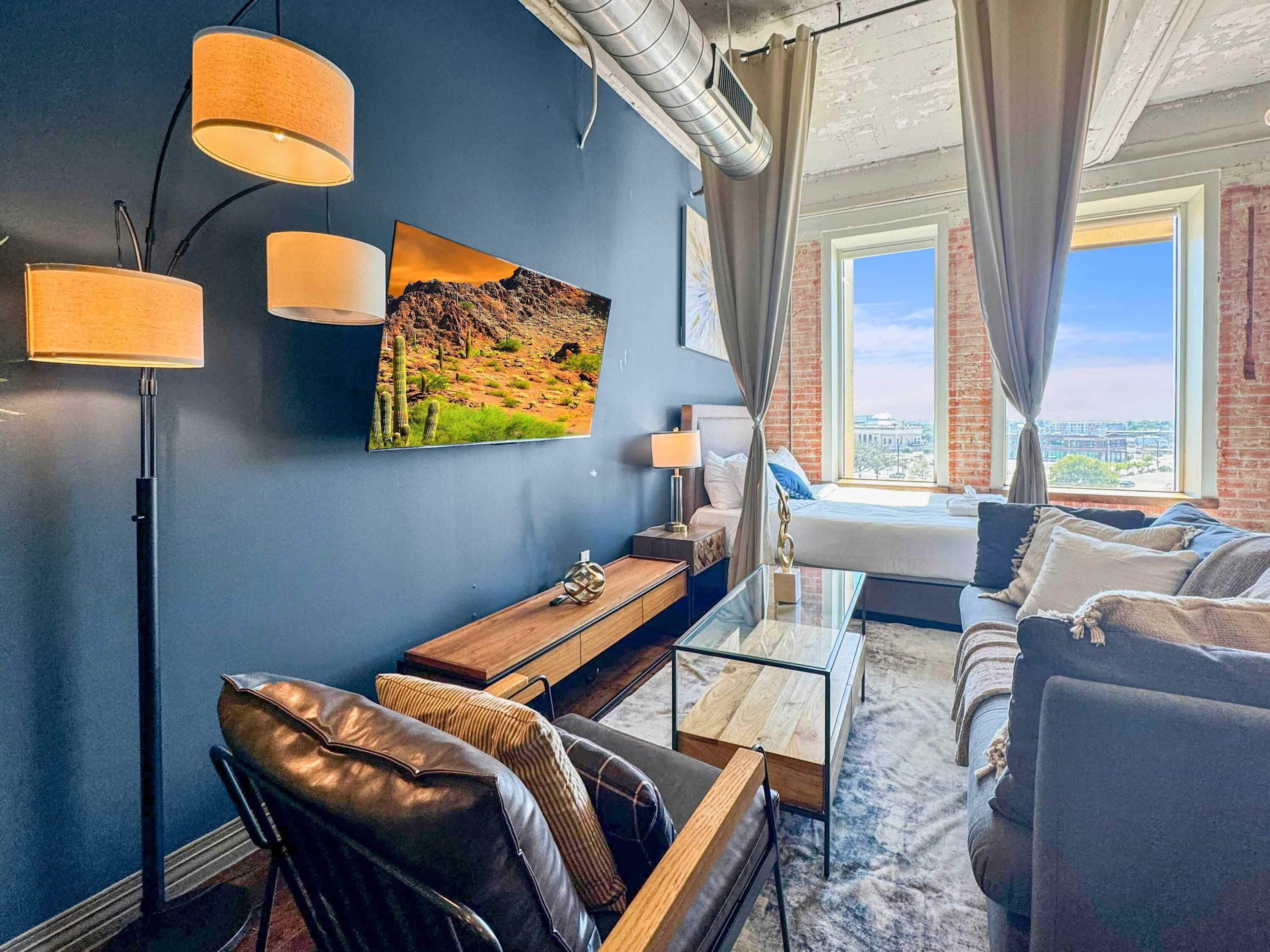 Una habitación de hotel moderna con una pared azul, un ventanal con vistas a la ciudad, una cama, una zona de estar con un sillón de cuero, una mesita de café y un cuadro de un paisaje desértico.