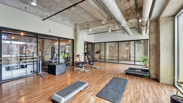 Un gimnasio con equipo de ejercicio, piso de madera y ventanas amplias.