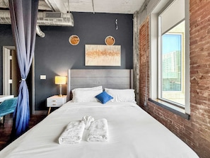 Una habitación de hotel con una cama, una mesita de noche, una lámpara, una ventana con vistas a edificios y una cortina.