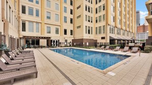 Zona de piscina de hotel con tumbonas, una barbacoa y un área de descanso techada.