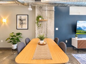 A conference room with a long table, chairs, a plant, a framed picture, a wall-mounted shelf with plants, and a television on the wall.