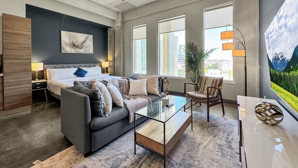 A modern hotel room with a bed, sofa, coffee table, and large windows offering city views.