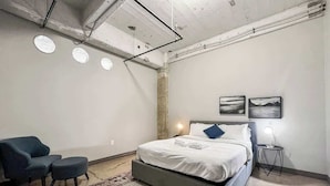 A modern bedroom with a large bed, a seating area with a chair and ottoman, a small side table, and a wall-mounted light.