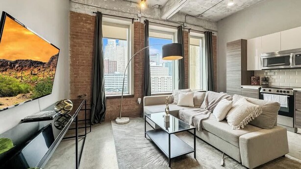 A modern living room with a sofa, coffee table, and TV. There are windows with curtains, a kitchen area with appliances, and a cityscape view.