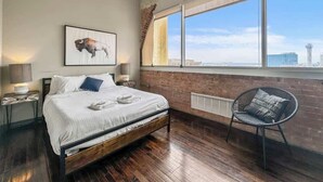 Ein modernes Schlafzimmer mit einem großen Bett, Blick auf die Stadt, einer Ziegelwand und einem Sessel am Fenster.