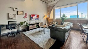 Ein modernes Wohnzimmer mit einer Ledersofa, einem Couchtisch und einem Fernseher. Es gibt einen Schreibtisch mit einem Stuhl, eine Pflanze und ein großes Fenster mit Blick auf die Stadt.