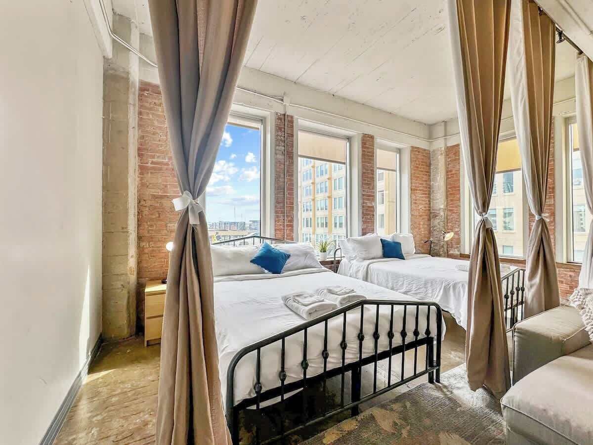 Een hotelkamer met twee bedden, een bakstenen muur, grote ramen en uitzicht op de stad.