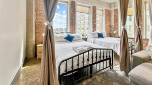 A hotel room with two beds, a brick wall, large windows, and a view of the city.