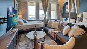 A modern hotel room with a bed, sofa, and a round table. There is a large window with curtains, a television, and a view of the city.