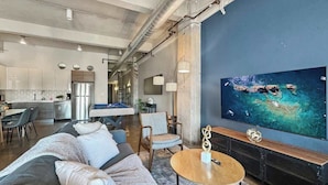 A modern living room with a large flat-screen TV, a sofa, a coffee table, and a kitchen in the background.