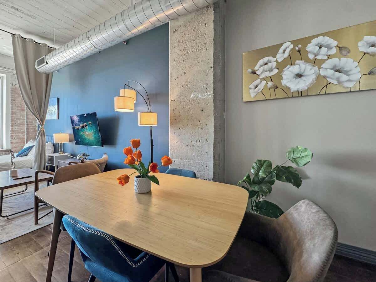 Un comedor moderno con una mesa de madera, sillas azules y un jarrón con flores naranjas. Un cuadro grande de flores blancas cuelga en la pared, y se observa una planta en la esquina.