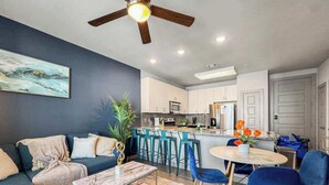 Una sala moderna con sofá, mesa de centro y sillones, un área de cocina con taburetes de bar y un ventilador de techo.