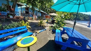 Um café à beira-mar com bancos azuis e uma mesa, rodeado por plantas em vasos e vista para o mar.