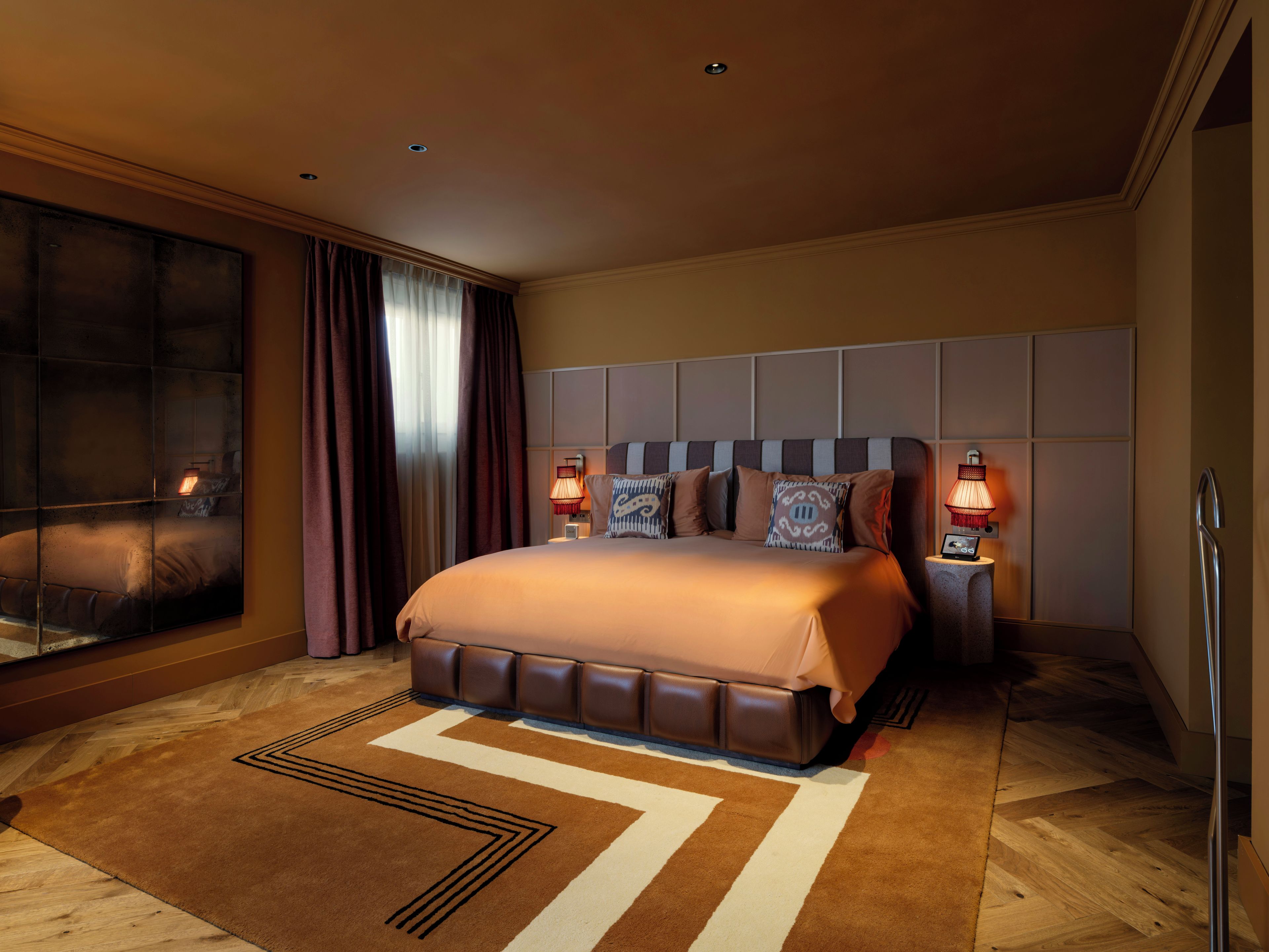 Habitación de hotel con una cama grande, dos lámparas de noche, un ventanal con cortinas y vistas a la ciudad.