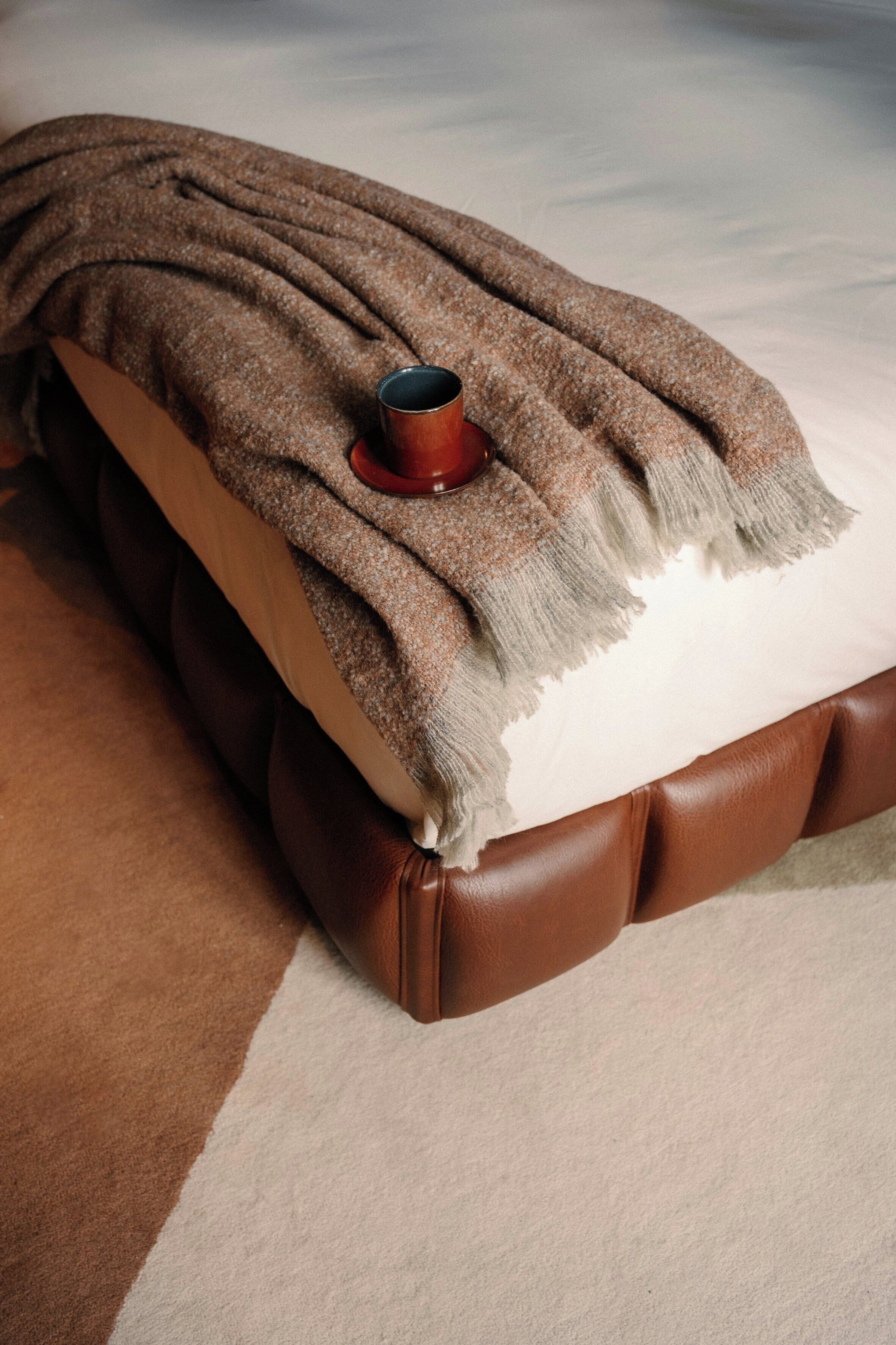 A bed with a brown leather headboard, a brown blanket, and a red cup on the bed.